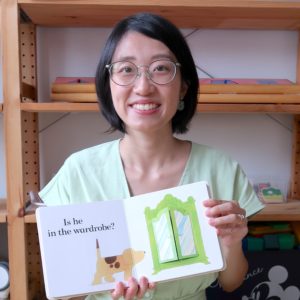 Sarah Tan, a woman with short hair and spectacles, is smiling and holding up a lift-the-flap book. The open book shows a dog looking into a green wardrobe.