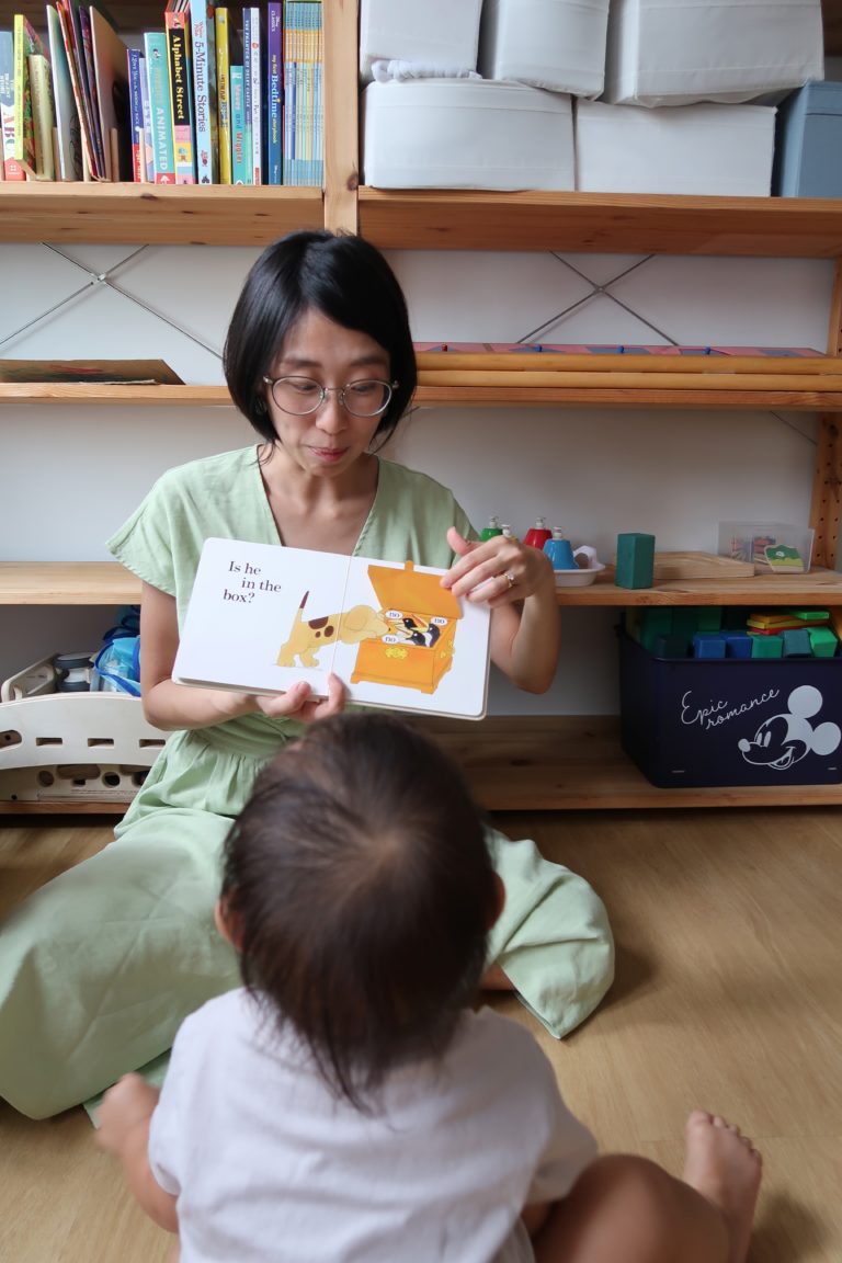 A woman in a green dress sits on the floor holding out a picture book, while a child, whose back faces the camera, watches her.