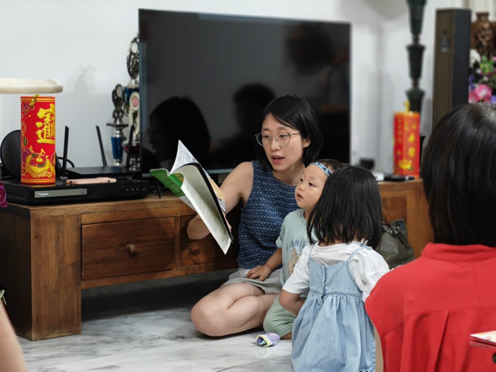 A woman sits on the floor with a child in her lap. She is holding a book and reading.