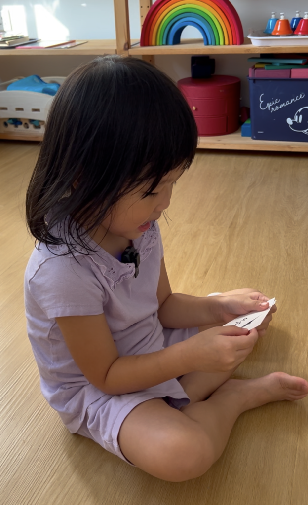 A girl sits holding a flashcard, which she is reading. Behind her is a shelf full of colourful toys.